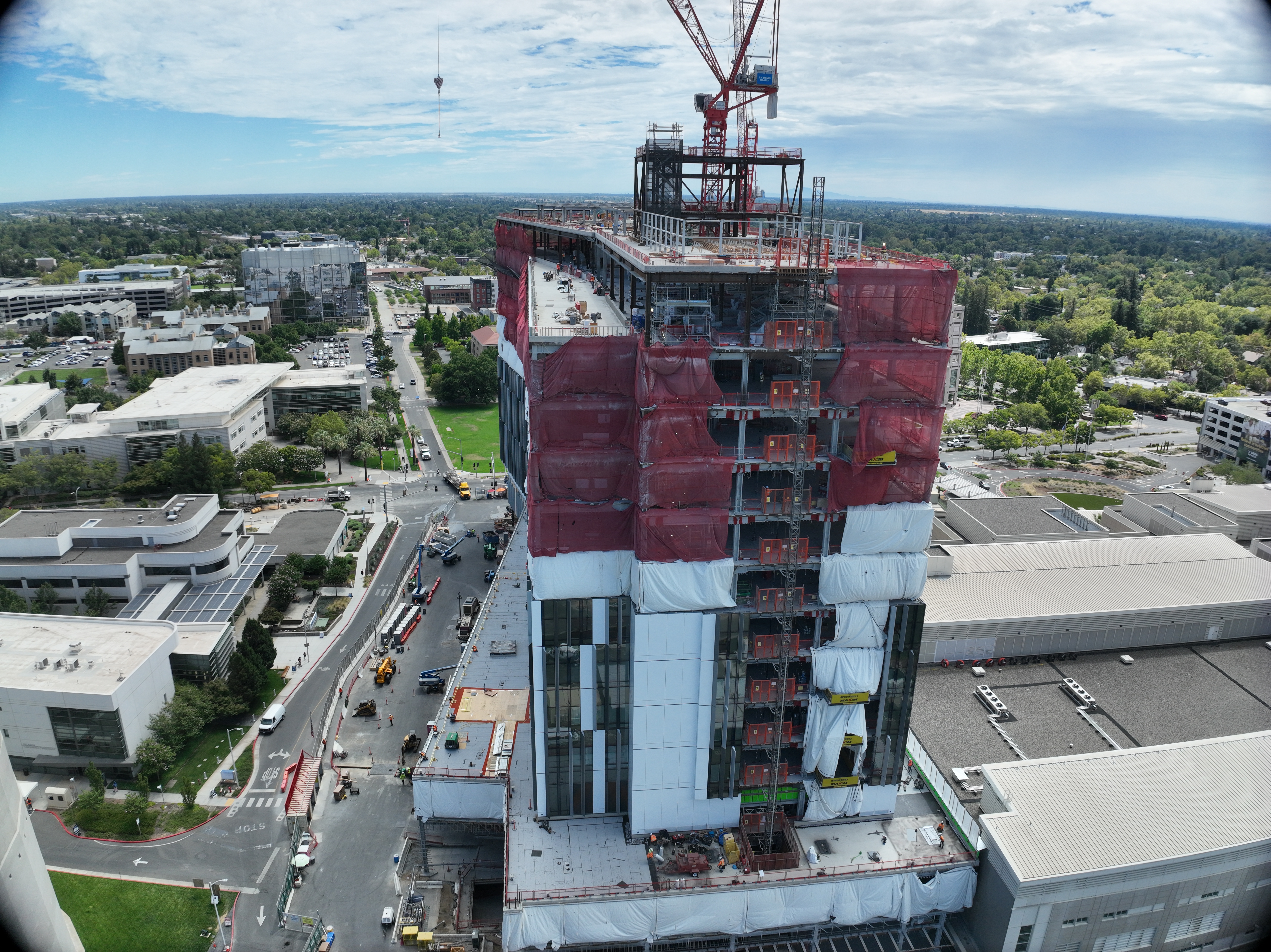 Aerial view of California Tower under construction with red safety netting, white exterior wrap and two cranes above the structure on the Sacramento campus.