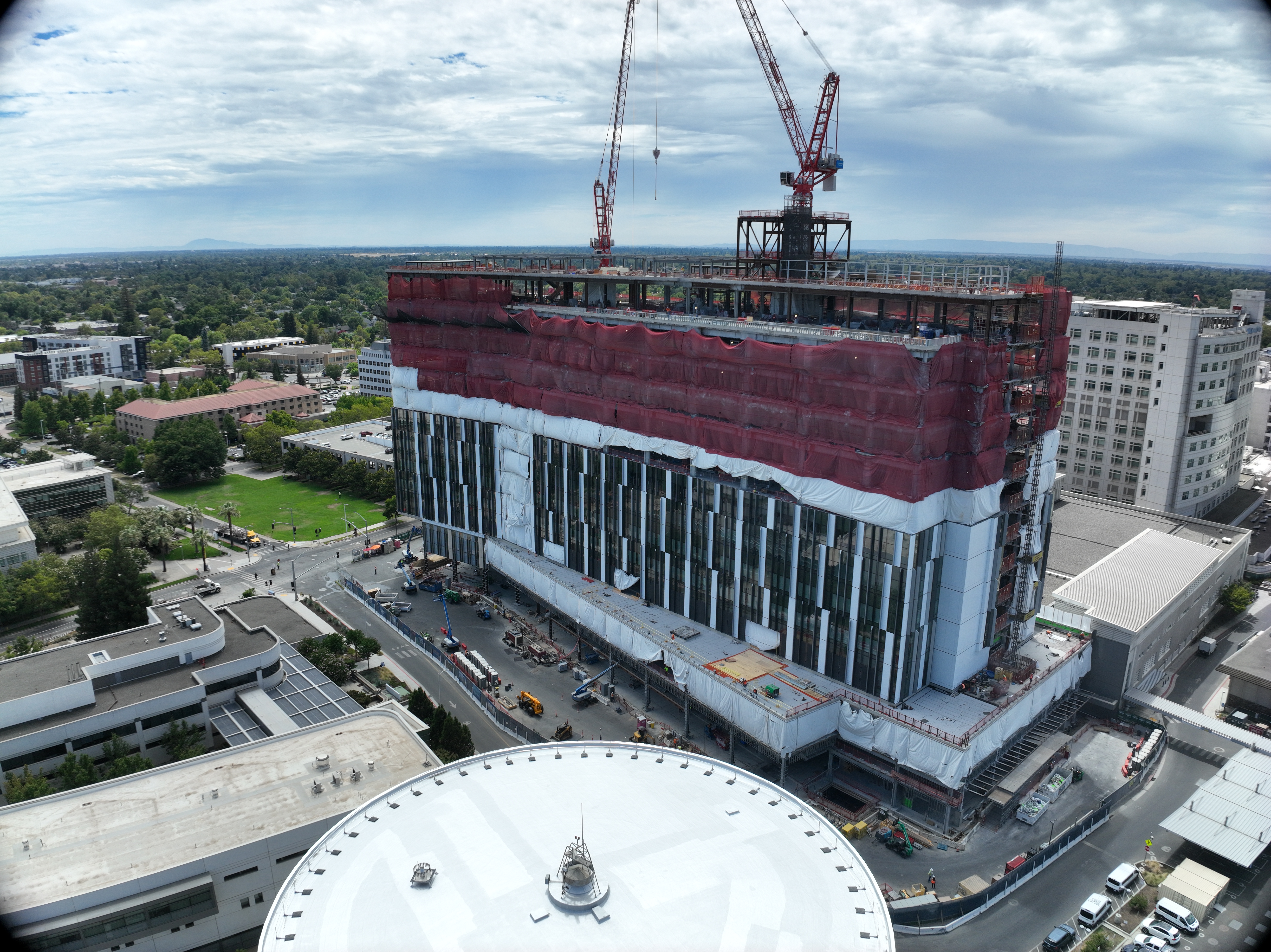 Aerial view of California Tower under construction with red safety netting, white exterior wrap and two cranes above the structure on the Sacramento campus.