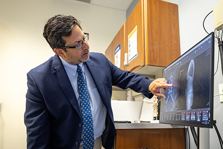 Man in dark blue suit points to two x-ray images on a computer screen. One image shows a close up of a spine with implants and screws. The other image is an opaque outline of a person’s back.