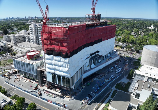 Wide aerial view of California Tower construction site with surrounding healthcare buildings, cranes overhead and red and white protective wrap.