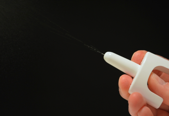 Close-up of a hand using a nasal spray device, with a fine mist spraying from the nozzle against a dark background.