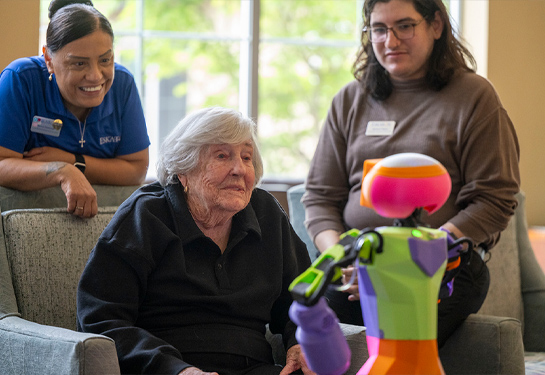 An older adult sits in a lounge watching a colorful robot, with two staff members nearby observing the interaction.