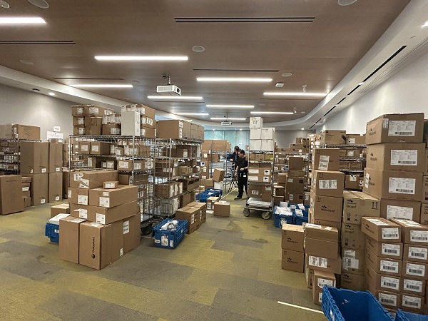 Wide view of a room filled with stacked boxes and shelving as staff organize supplies for distribution.