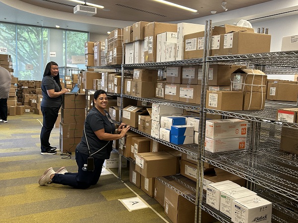 Two staff members sort and organize boxes of medical supplies on metal shelving inside the distribution hub.