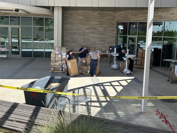 Staff members stand near stacks of boxes and pallets in an outdoor courtyard with equipment and carts nearby.