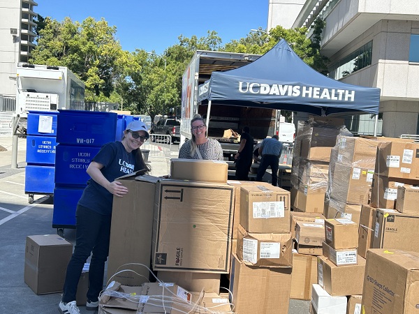 Two staff members in UC Davis Health shirts stand near an open truck and stacks of boxes while organizing incoming supplies.