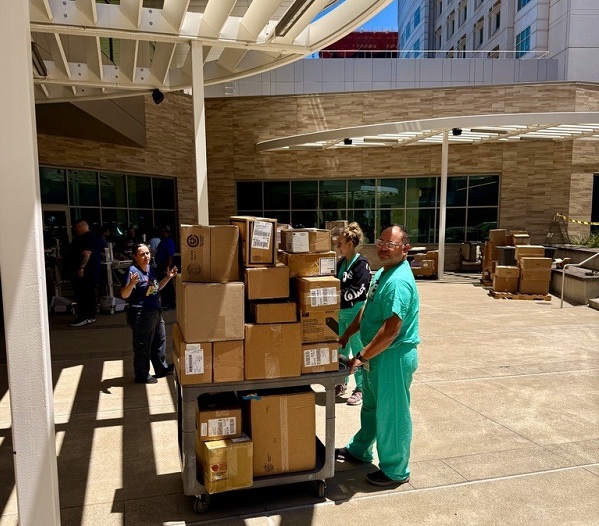 Staff pushes a cart stacked with boxes through an outdoor courtyard with additional supplies staged nearby.