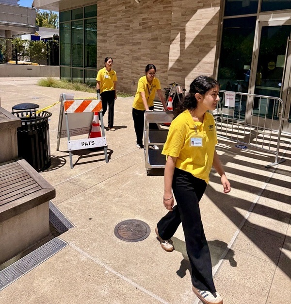 Volunteers in yellow shirts push a cart of supplies along an outdoor walkway near campus buildings.