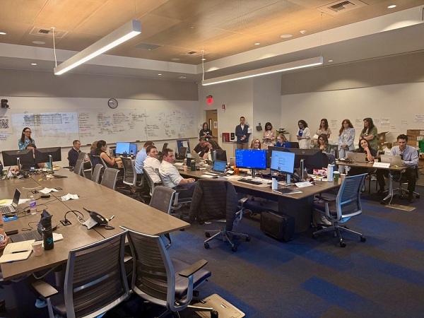 Room with multiple staff seated at desks and standing near whiteboards filled with notes while reviewing information on computers in a Command Center setting