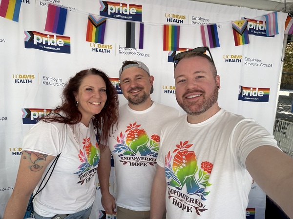 Three people in white Empowered Hope shirts take a selfie in front of a UC Davis Health Pride backdrop.