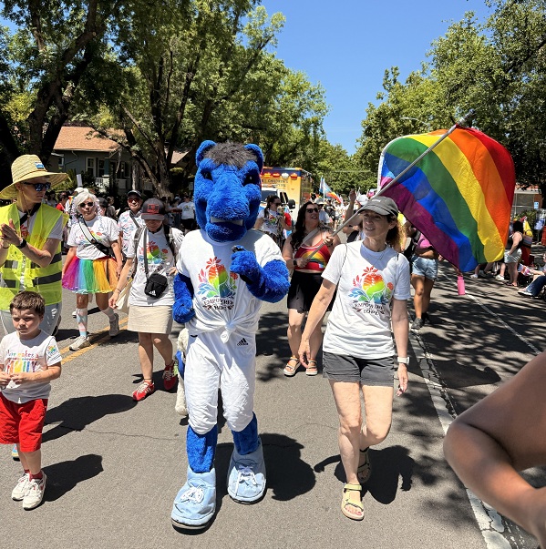 Blue mascot in white uniform walking in a parade next to a person holding a rainbow flag.