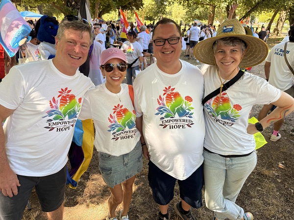 Four people in matching Empowered Hope shirts stand together at a Pride event with flags in the background.