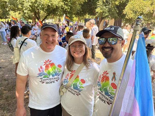 Three people in Empowered Hope shirts standing together outdoors, one holding a transgender pride flag.