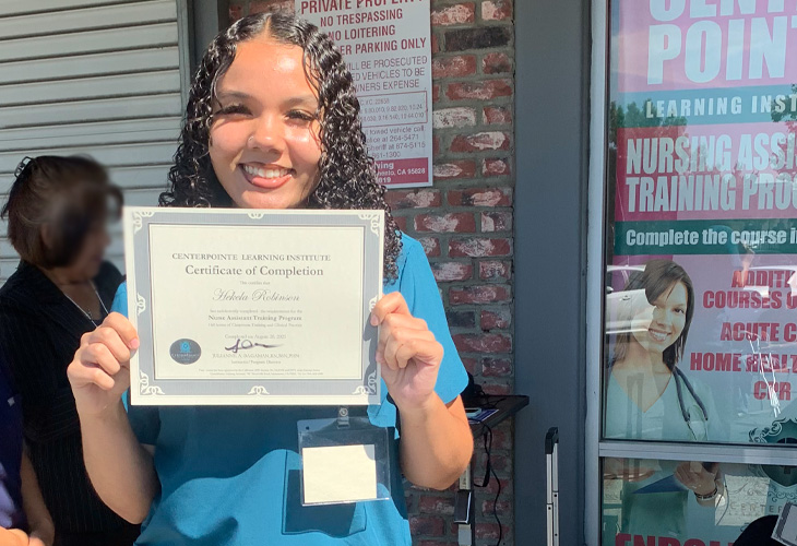 Person in scrubs smiles while holding a certificate of completion for a nurse assistant training program