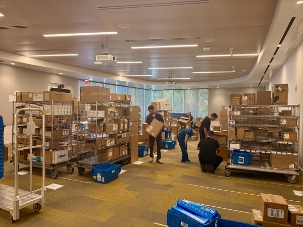 Indoor room with shelving units and carts filled with boxes and supplies as several people sort and organize inventory for distribution