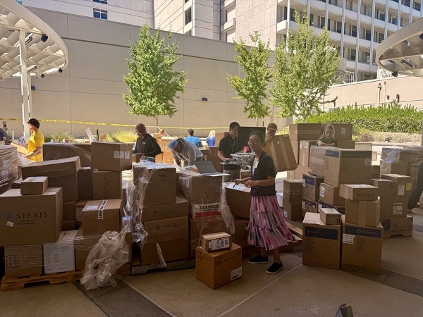 Boxes of medical supplies arranged on the ground and on pallets as several people sort and handle items in an outdoor staging area