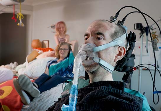 A man in a wheelchair with a breathing tube and cables attached to his head for a brain-computer interface sits in his living room. 