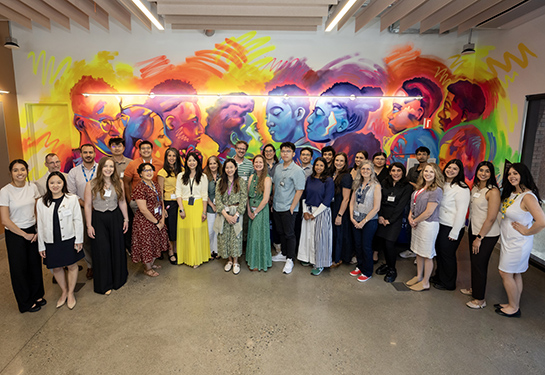 Large group of people standing in front of a colorful mural with light shining above them. 