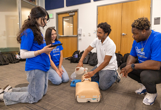 Student participants practice emergency response skills using a CPR mannequin lying on floor during a hands-on training session