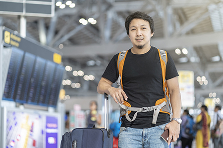 Man wearing backpack and holding passport standing next to flight information screen at airport