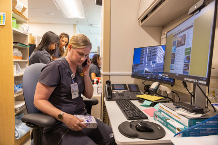 A healthcare worker before computer, talking on phone and reviewing a medication package, with colleagues in the background.