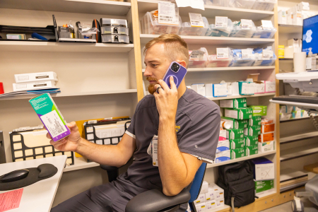 A healthcare worker sits in a supply room, reviewing a medication package while talking on the phone.