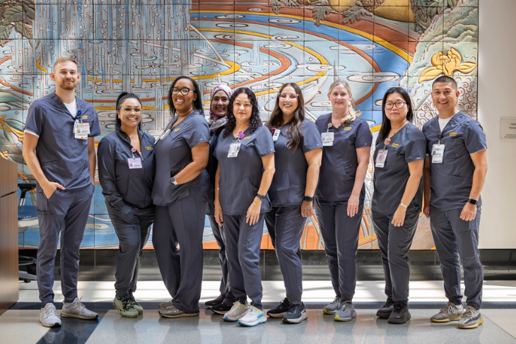 A group of healthcare team members in matching scrubs pose together in front of a colorful wall mural.