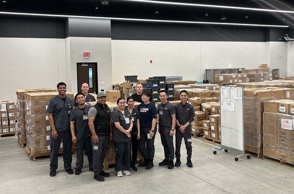 Employees stand in a warehouse area with pallets of boxed medical supplies staged throughout the room.
