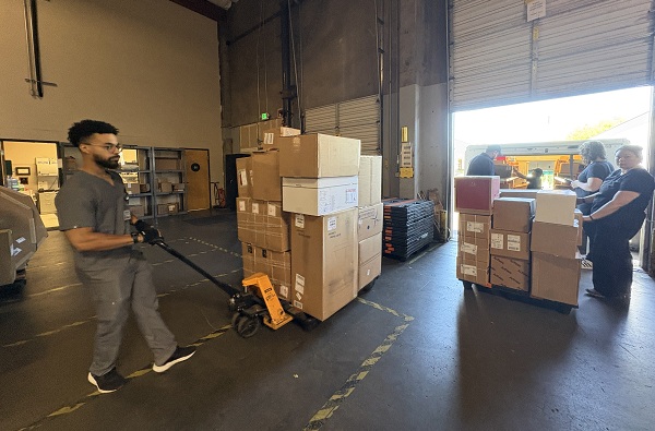 Employee moves a pallet loaded with boxes through a warehouse loading area while additional shipments are staged nearby.