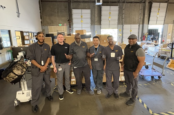 Six employees stand in front of pallets and boxes inside a warehouse.