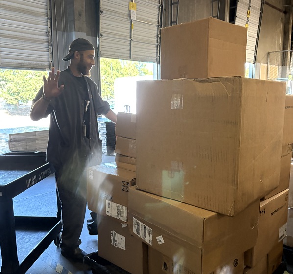 Employee stands beside a cart loaded with stacked cardboard boxes near a warehouse loading door.