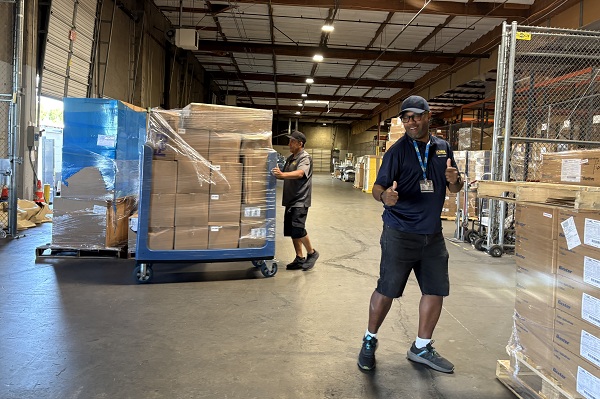 Employee walks through a warehouse aisle while a large cart loaded with supplies is pushed in the background.
