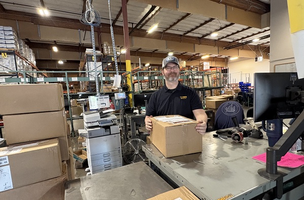 Employee stands at a workstation holding a box in a warehouse filled with shelving and inventory.