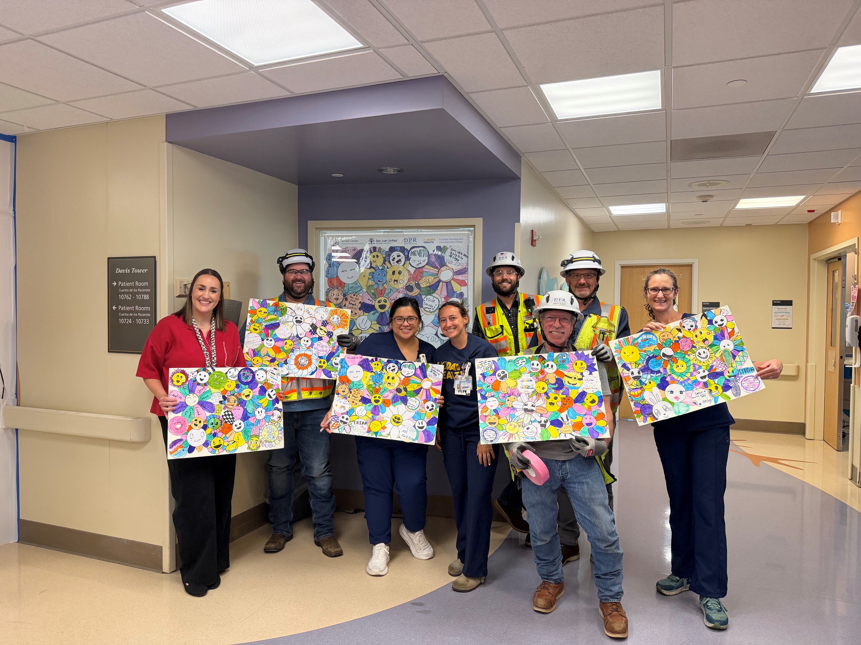 Construction workers, hospital staff and nurses pose in a Pediatric Intensive Care Unit hallway holding colorful student artwork that is displayed on construction barriers behind them.