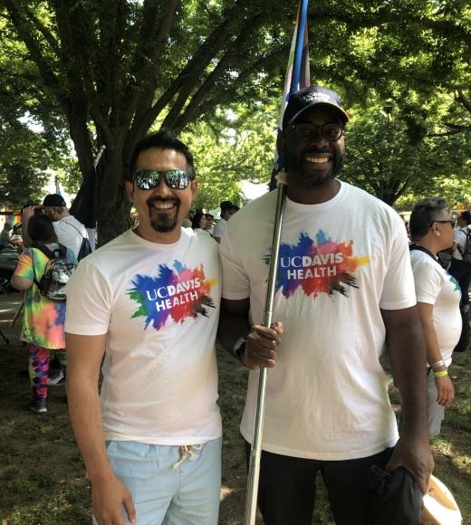 Two people wearing UC Davis Health Pride shirts stand outdoors among event participants and trees.