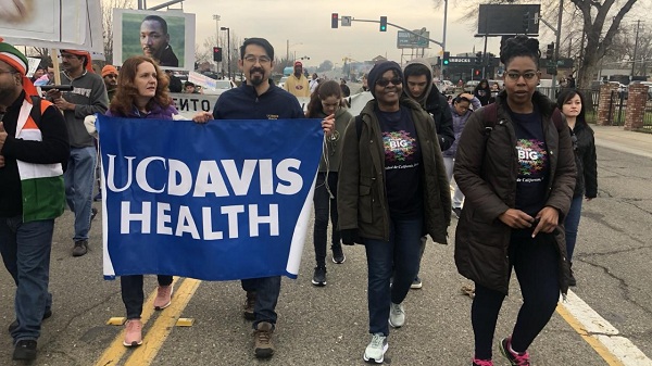 A group of people walk down a city street carrying a UC Davis Health banner during a community march.
