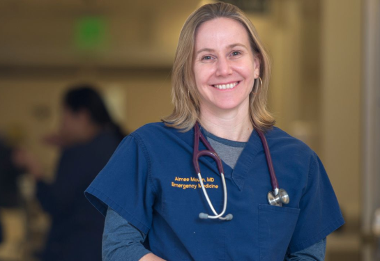 A person wearing blue medical scrubs with a stethoscope stands in sharp focus in a healthcare setting.