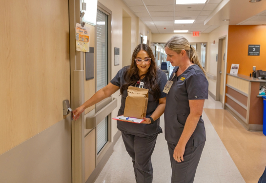 Two healthcare workers walk through a hospital hallway carrying a clipboard and a paper bag while opening a patient room door.