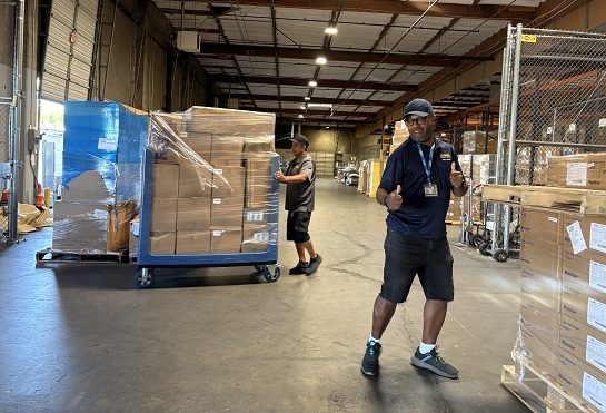 Employee walks through a warehouse aisle while a large cart loaded with supplies is pushed in the background.
