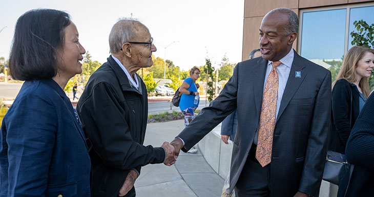 A woman in a blue jacket stands next to an older man who is shaking hands with a man in a blue suit. 