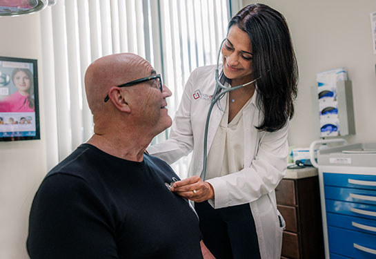 A physician in a white coat leans over a seated, older man while examining him.