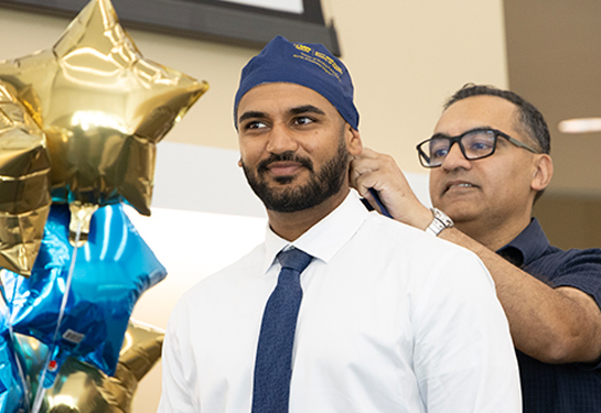 A man adjusts a nurse anesthesia student’s blue surgical cap during a capping ceremony, with blue and gold star-shaped balloons in the background.