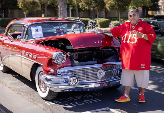 Car show attendee poses beside a red-and-white classic Buick with the hood open at a car show.