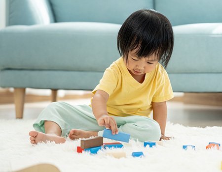 Toddler with dark hair, yellow shirt and green pants plays on a rug with colorful wooden blocks.