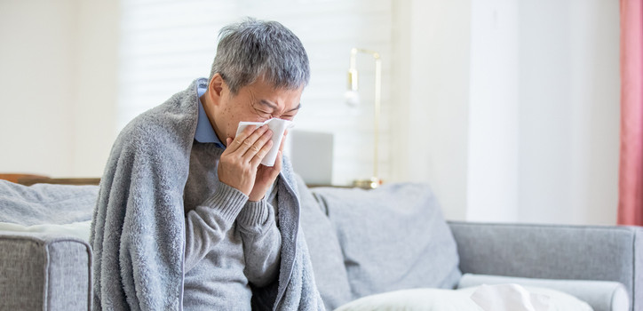 A man sits on a couch with a blanket and a tissue to his nose