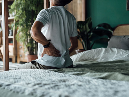 Rear view of senior Asian man suffering from backache, massaging aching muscles while sitting on bed.