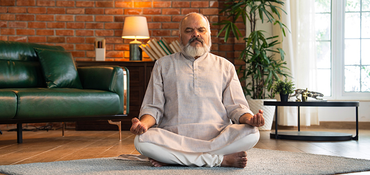 A senior man with his eyes closed sits on a carpet practicing deep breathing in a modern home.