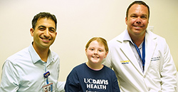 From left to right, Dr. Cameron Sadegh, Emily Roberts and Dr. Rick Price sit on an exam room bed. The doctors wear white coats, and Emily wears a navy blue t-shirt with text that reads UC Davis Health Department of Neurological Surgery.