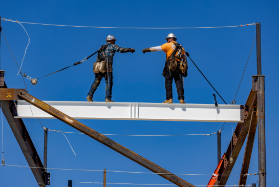 two construction workers standing on a steel beam in the air and fistbumping each other