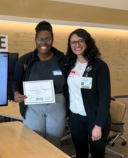 Two people pose, both wearing dark jackets and glasses, as one holds a certificate recognizing an award.